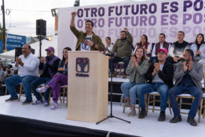 Pedro Kumamoto está en el estrado, levanta su brazo derecho con el puño cerrado para demostrar la fuerza de su movimiento. Detrás de él se encuentran algunos otros actores y actrices políticas. Se puede leer en una pancarta grande la frase: Otro Futuro Es Posible